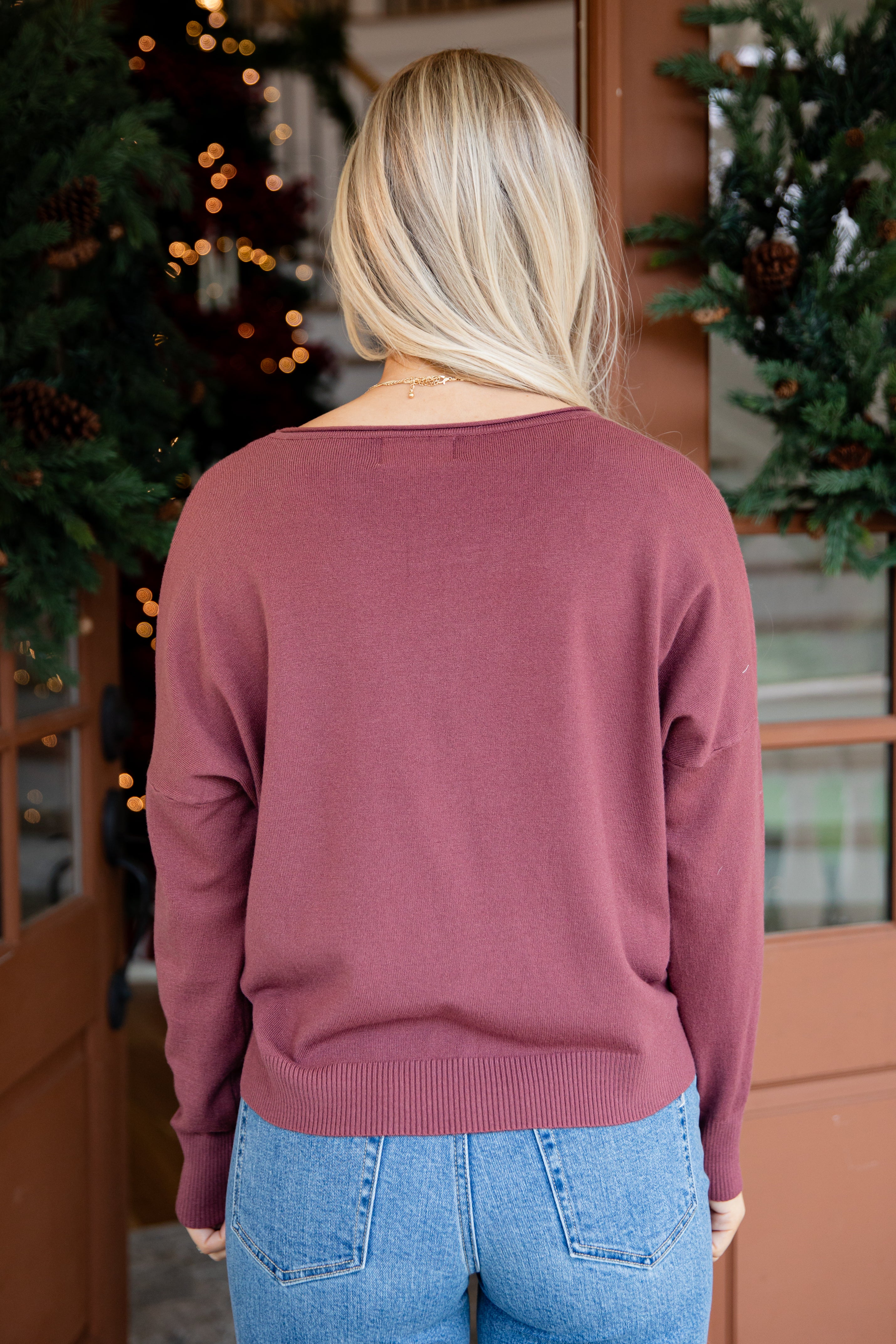 Person wearing a mauve sweater and blue jeans standing in front of Christmas trees.