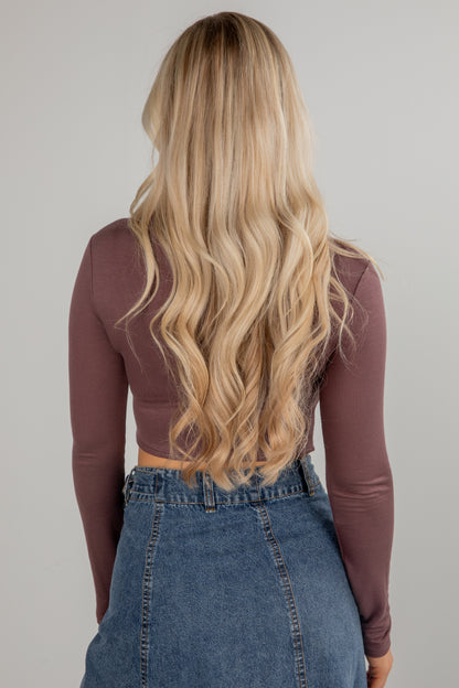 Person with long blonde hair wearing a brown long-sleeve top and blue jeans on a plain background