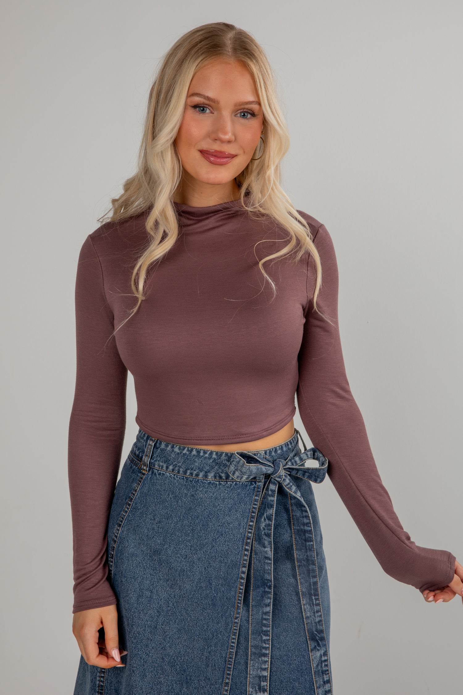 Woman wearing a brown long-sleeve top and blue denim skirt against a gray background