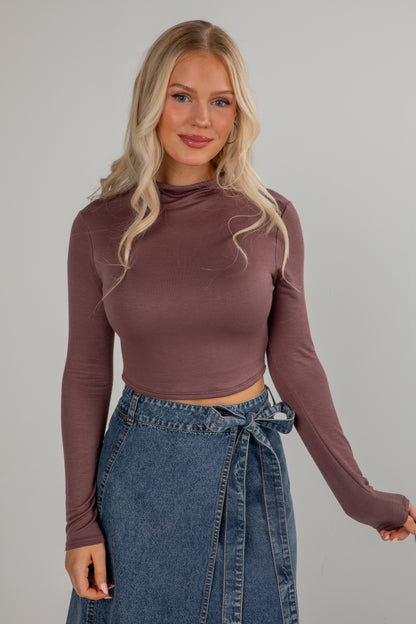 Woman wearing a brown long-sleeve top and blue denim skirt against a gray background