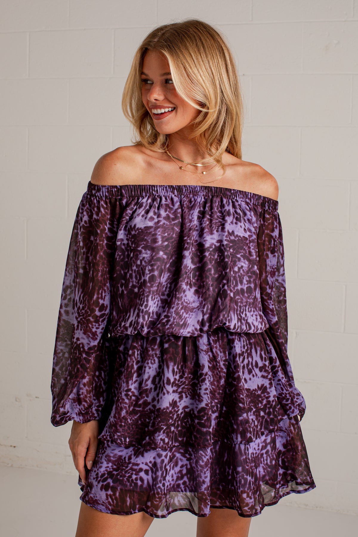 A woman wearing a purple and lavender abstract patterned off-the-shoulder mini dress with long sheer sleeves, standing against a white painted brick wall.
