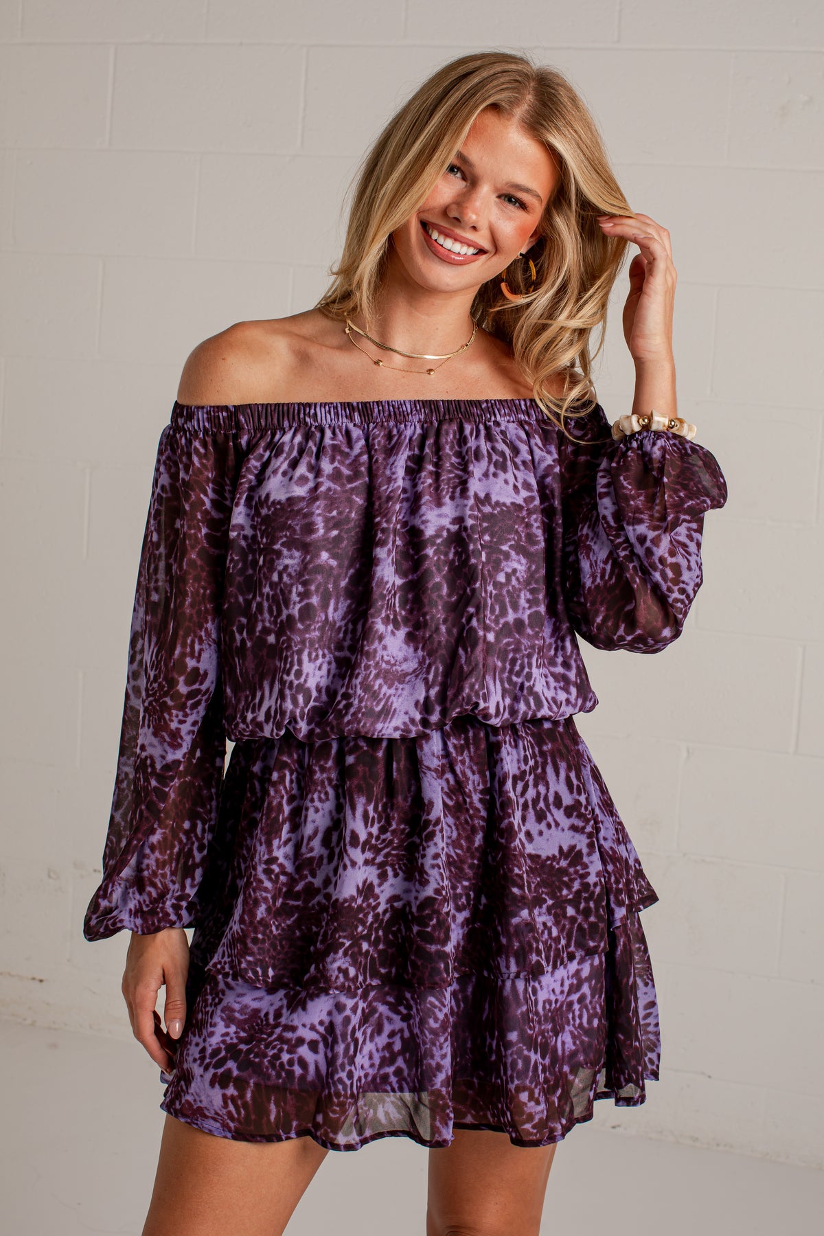 A woman modeling an off-the-shoulder purple and black patterned mini dress with long puff sleeves, set against a light gray brick wall.