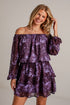 A woman modeling an off-the-shoulder purple and black patterned mini dress with long puff sleeves, set against a light gray brick wall.