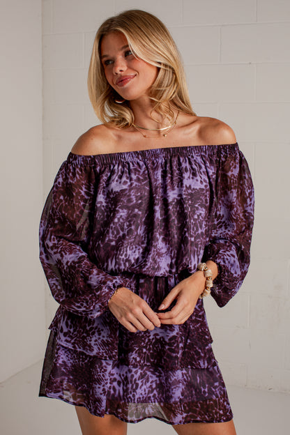 A woman wearing a purple off-the-shoulder long-sleeve mini dress with an abstract animal print pattern, standing against a white brick wall.