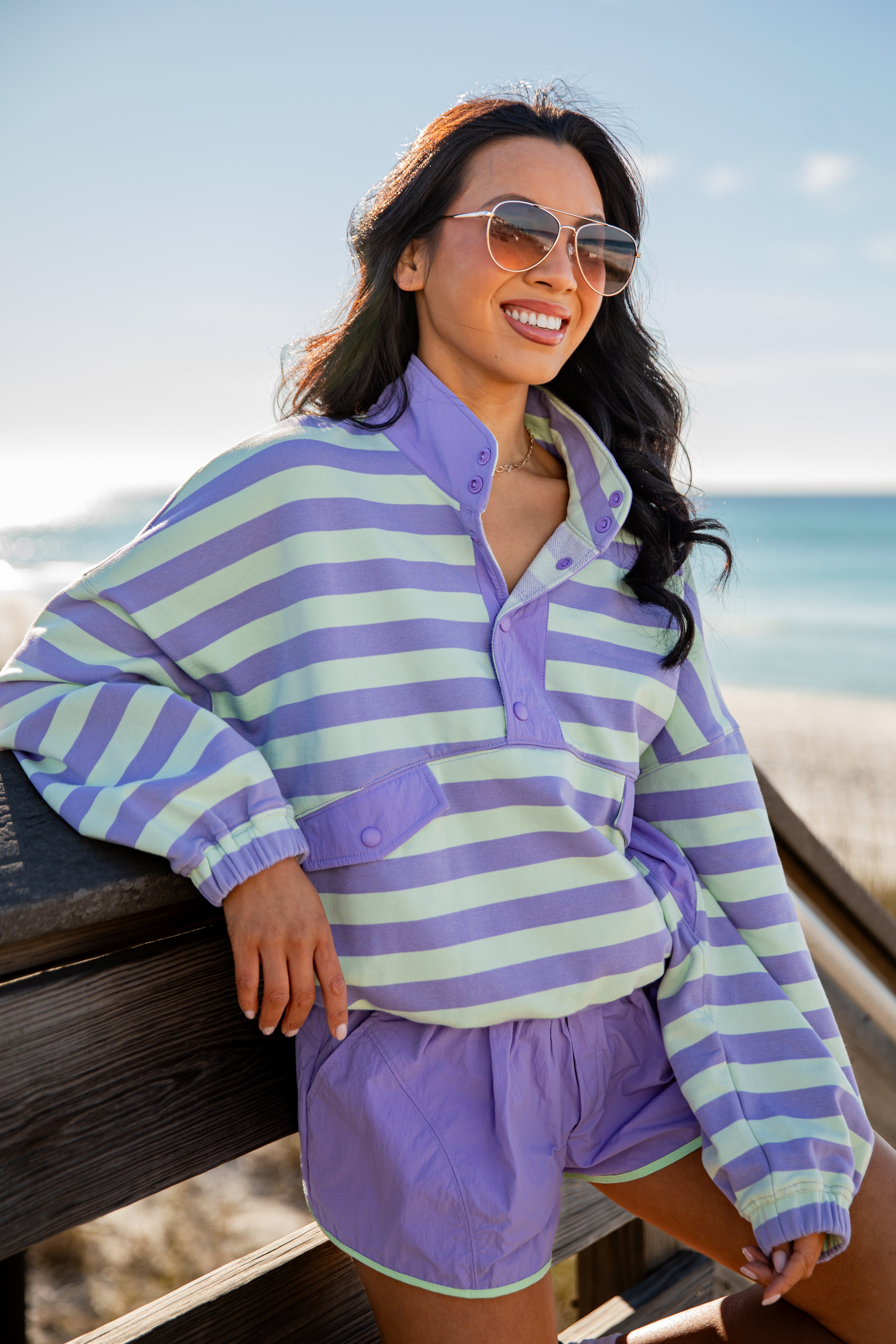 Woman wearing a purple and green striped shirt and shorts, sitting on a wooden bench by the beach.