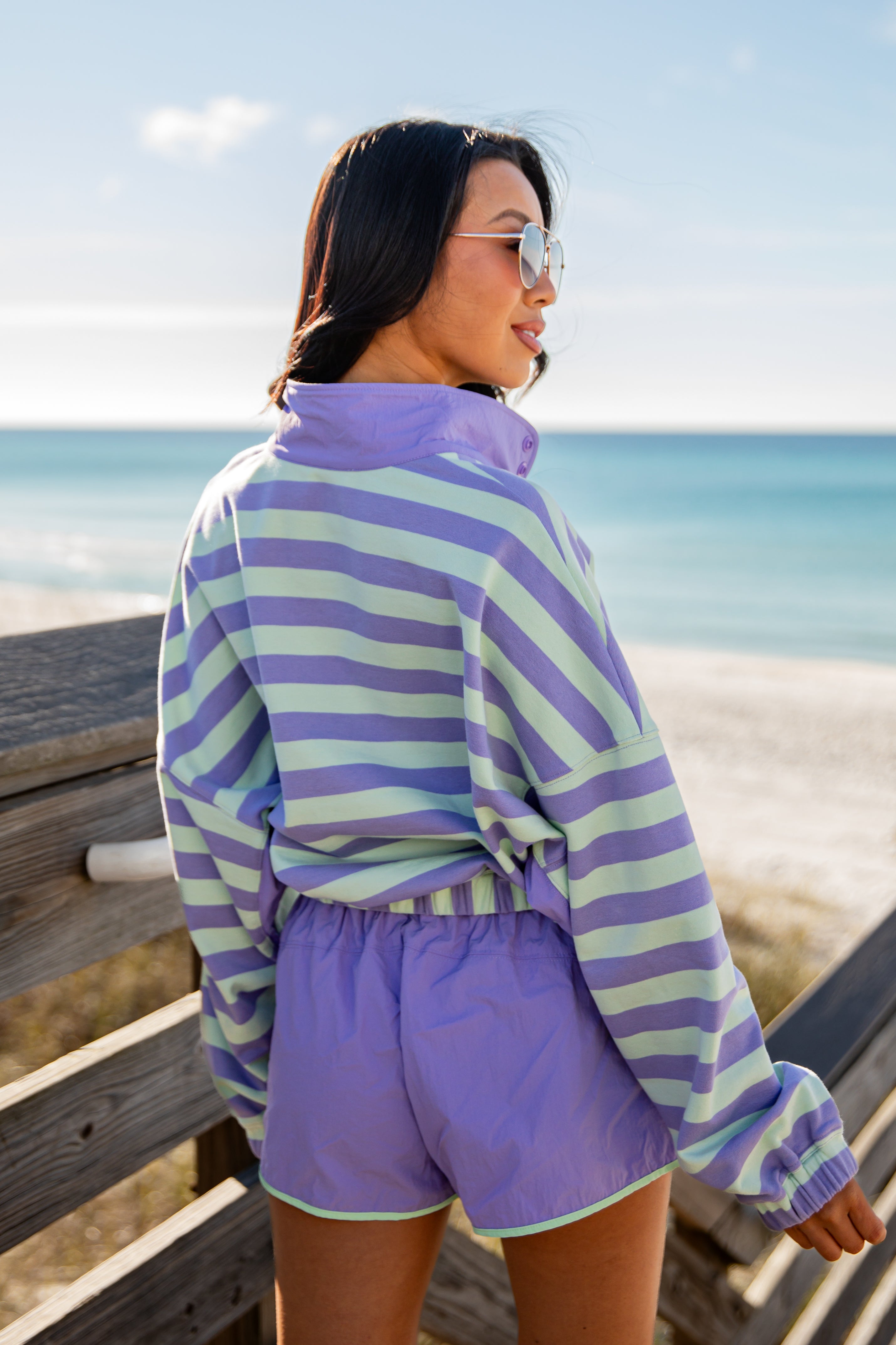 Woman wearing a purple and green striped shirt and shorts on a beach.