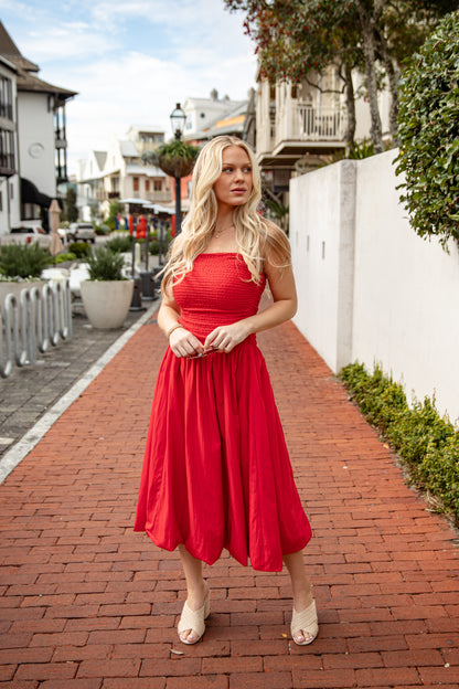 Woman in a red dress standing on a brick path with buildings and greenery in the background