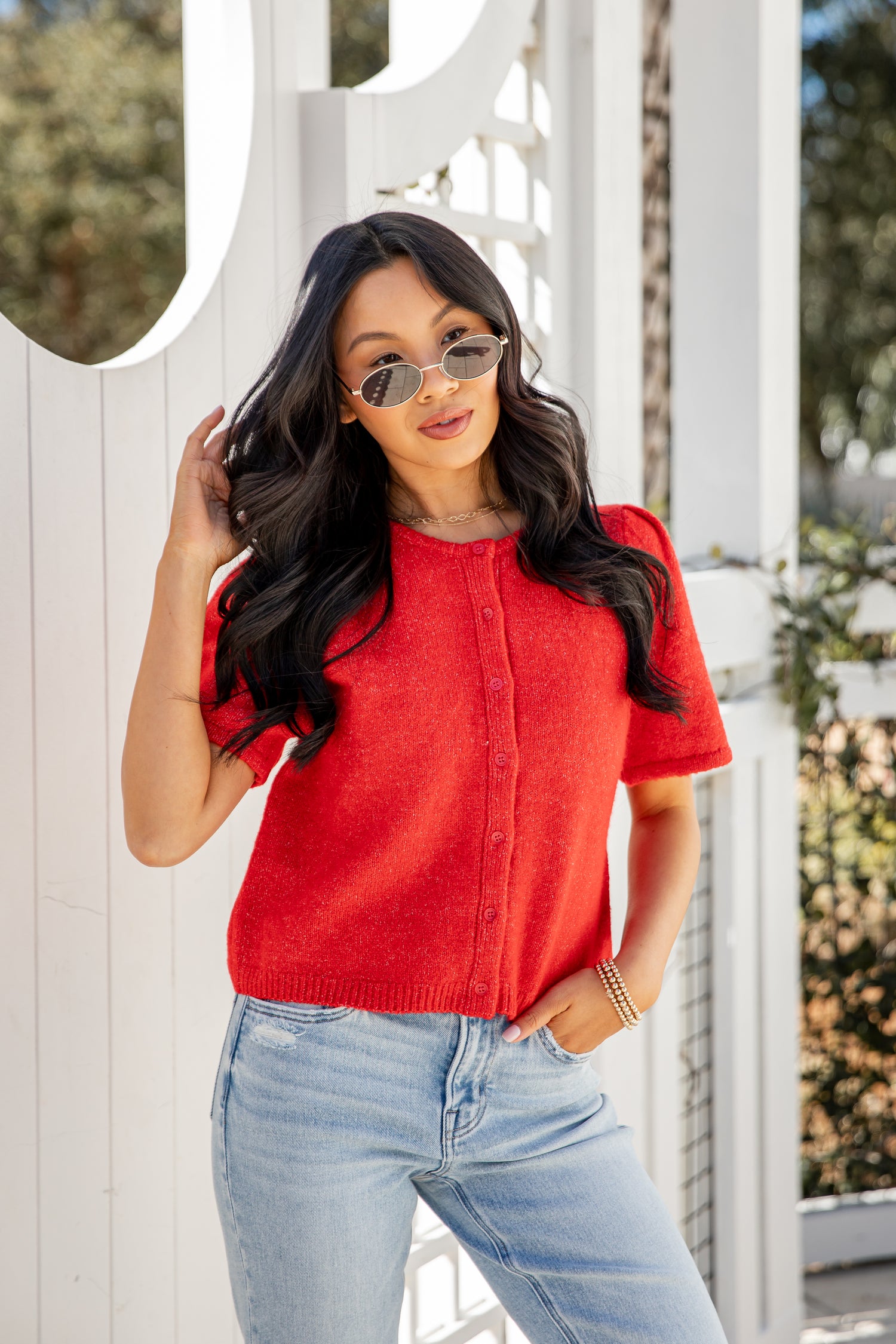 Woman wearing a red top and light blue jeans standing in front of a white gate.