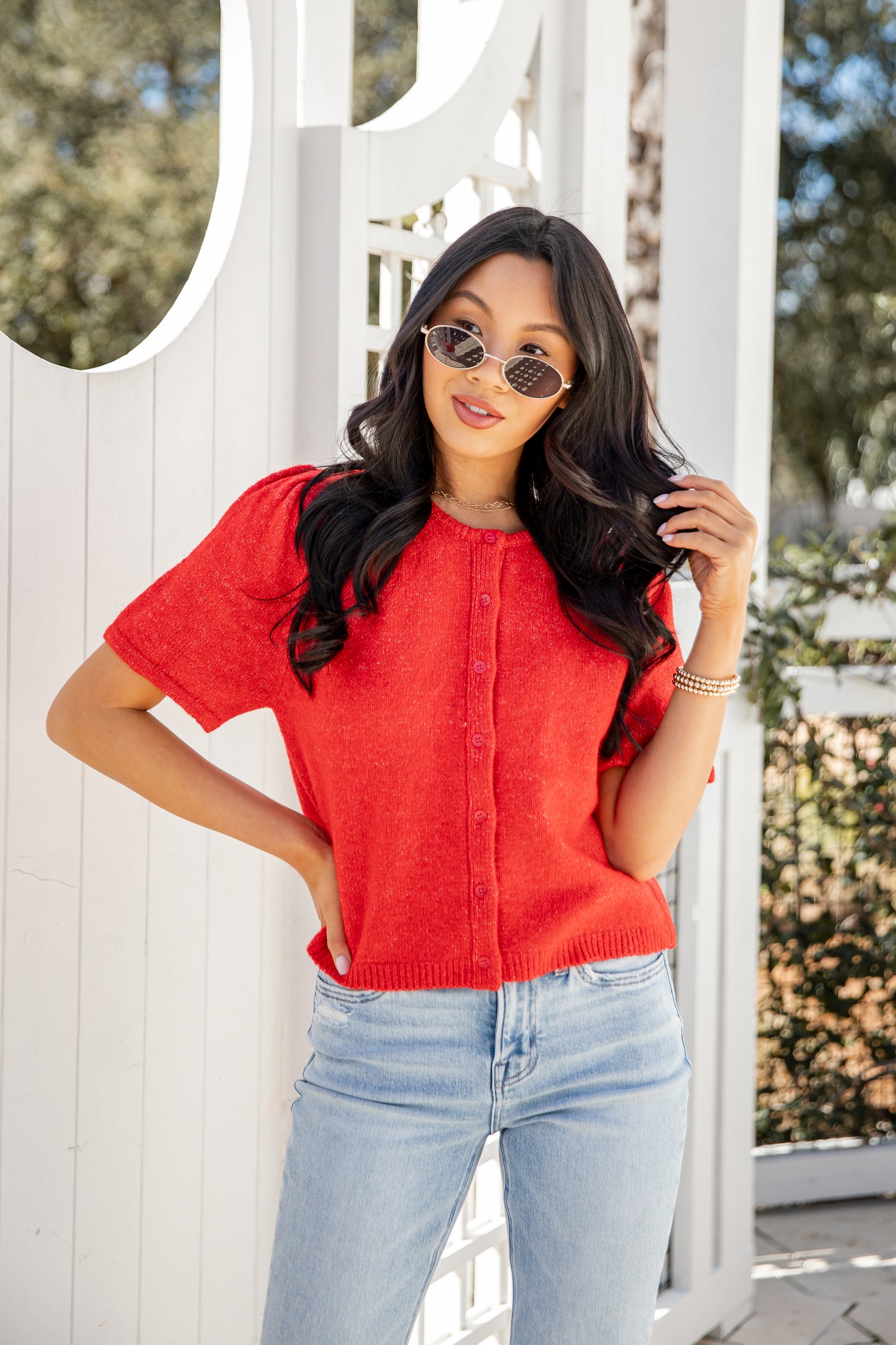 Woman wearing a red shirt and light blue jeans standing in front of a white wooden fence.