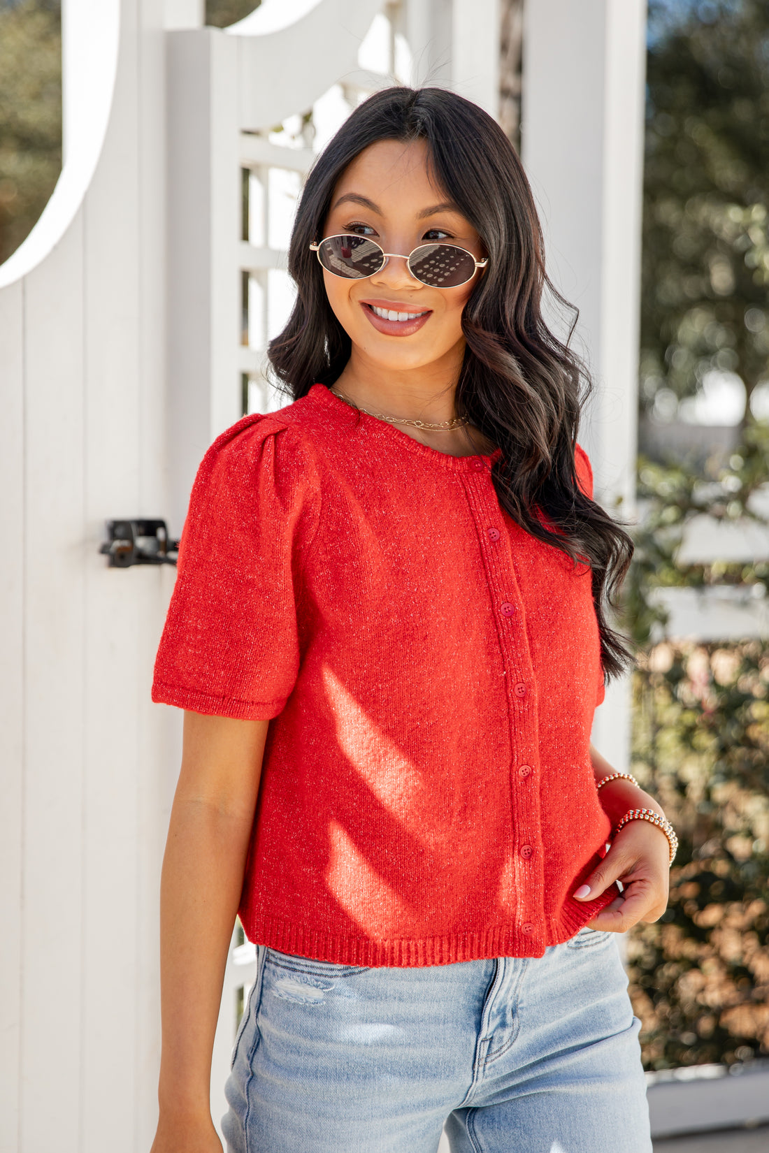 Woman wearing a red top and sunglasses, standing in front of a white door.