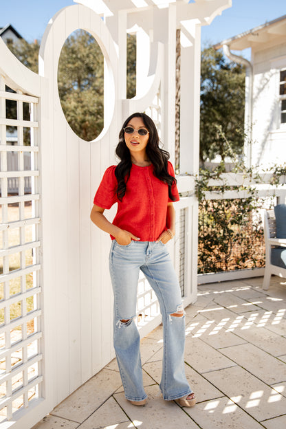 Woman in a red shirt and light blue jeans standing in front of a white lattice structure.