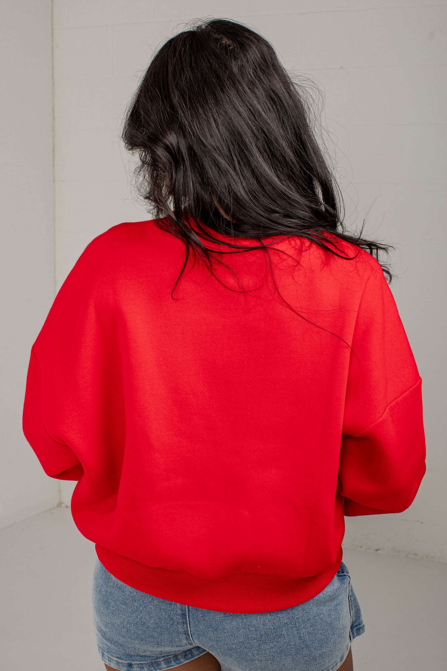 A person viewed from behind wearing a bright red crewneck sweatshirt and light blue denim shorts against a plain off-white wall.