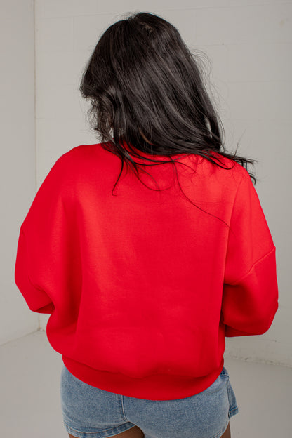 A person viewed from behind wearing a bright red crewneck sweatshirt and light blue denim shorts against a plain off-white wall.