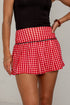 A close-up of a model wearing a red and white gingham pleated tennis skirt with a black waistband trim, paired with a black sleeveless top.
