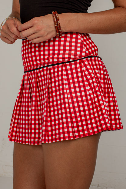A red and white gingham pleated tennis skirt with a thin black waistband, shown on a model against a light gray background.
