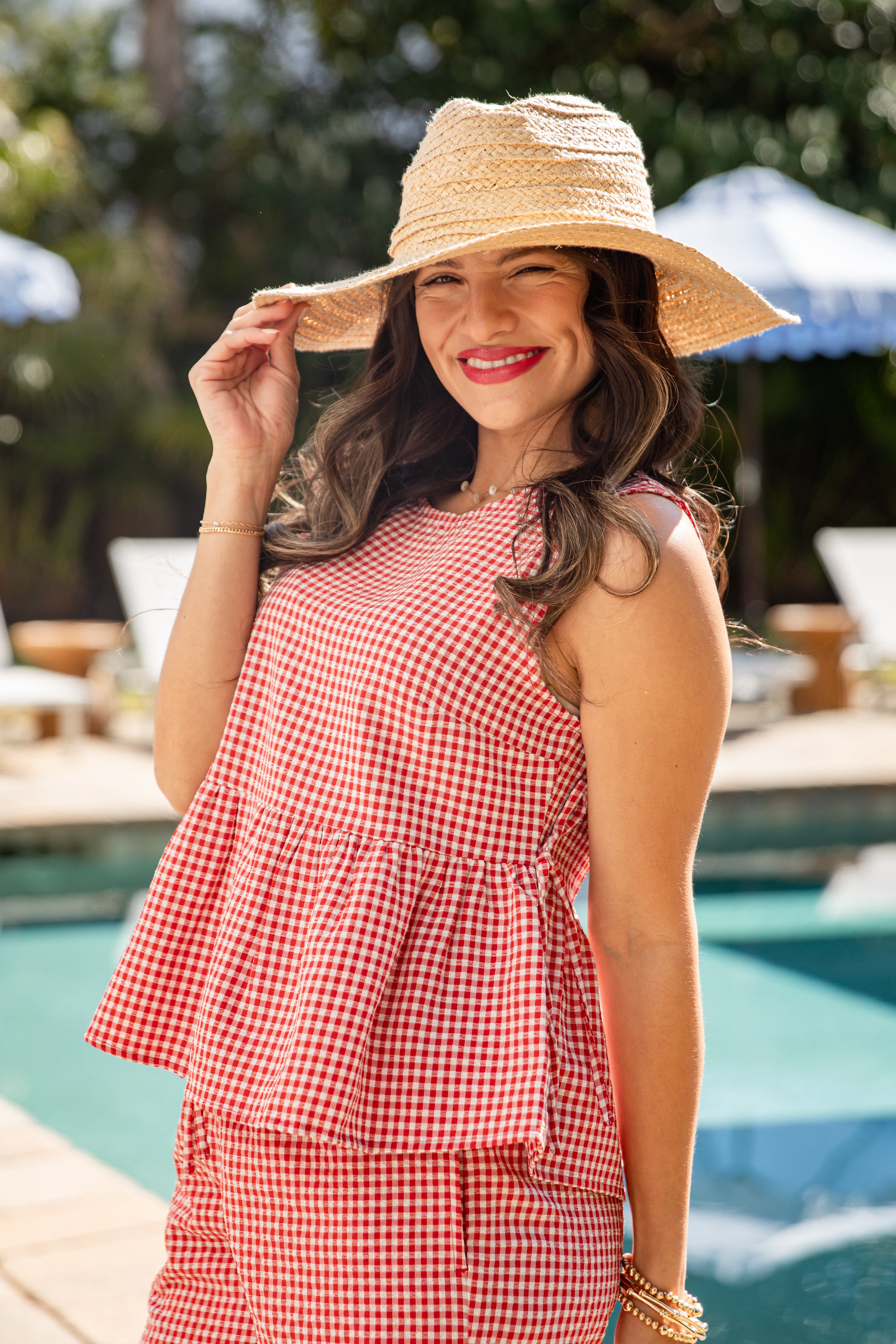 Woman wearing a red checkered dress and straw hat by a poolside.