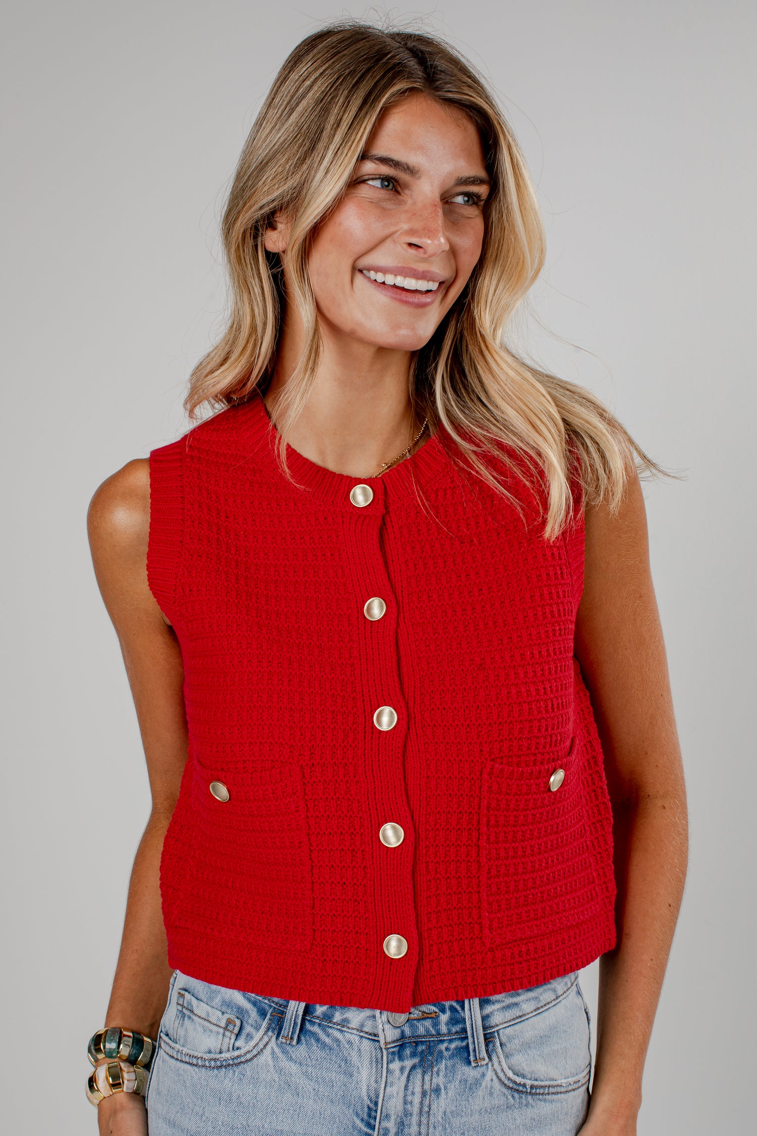 A woman wearing a bright red sleeveless cropped knit vest with gold-tone buttons and front pockets, paired with light-wash blue jeans, against a solid light gray studio background.