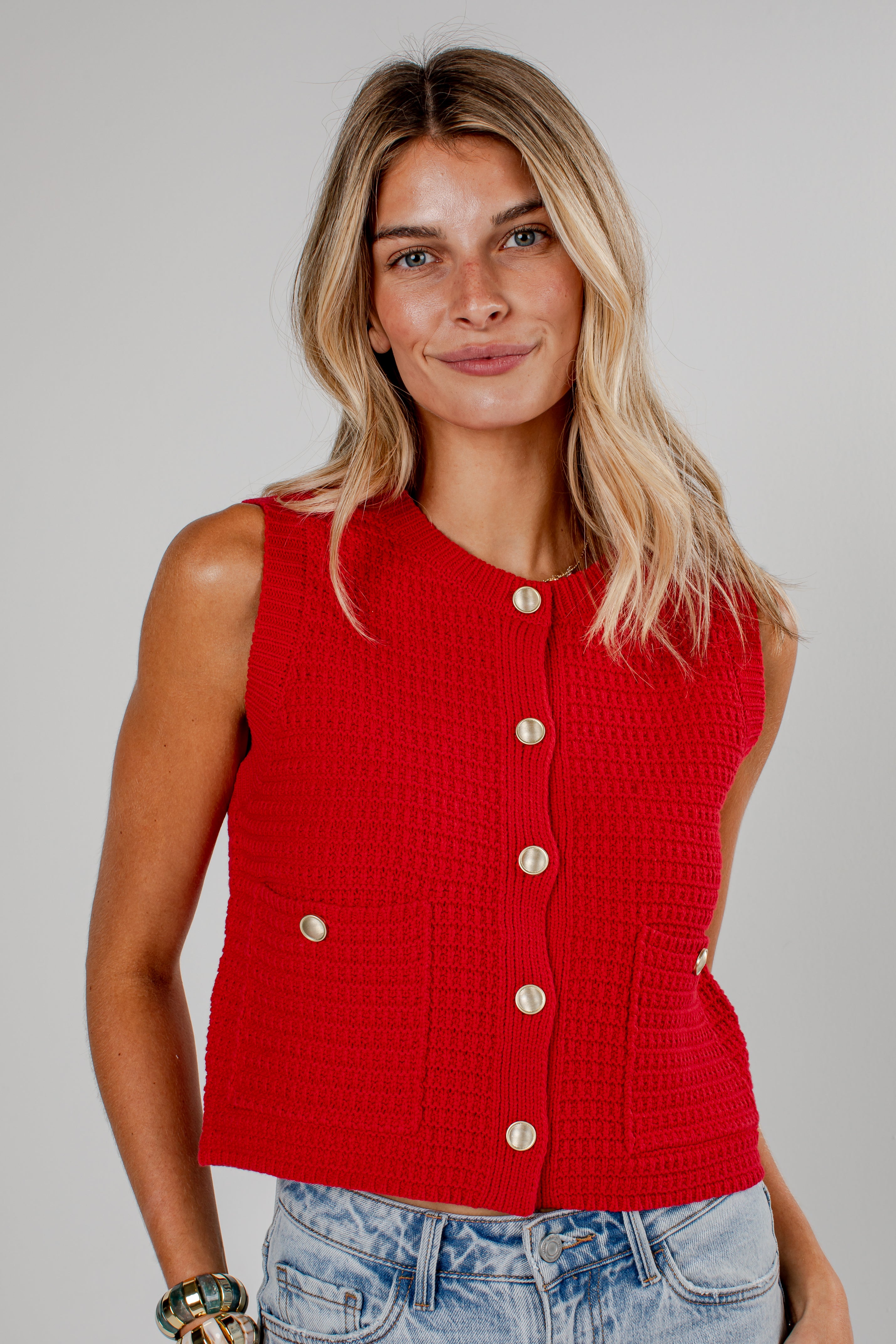 A woman wearing a bright red sleeveless textured knit vest with gold-tone buttons, paired with light-wash blue jeans, against a solid light gray studio background.