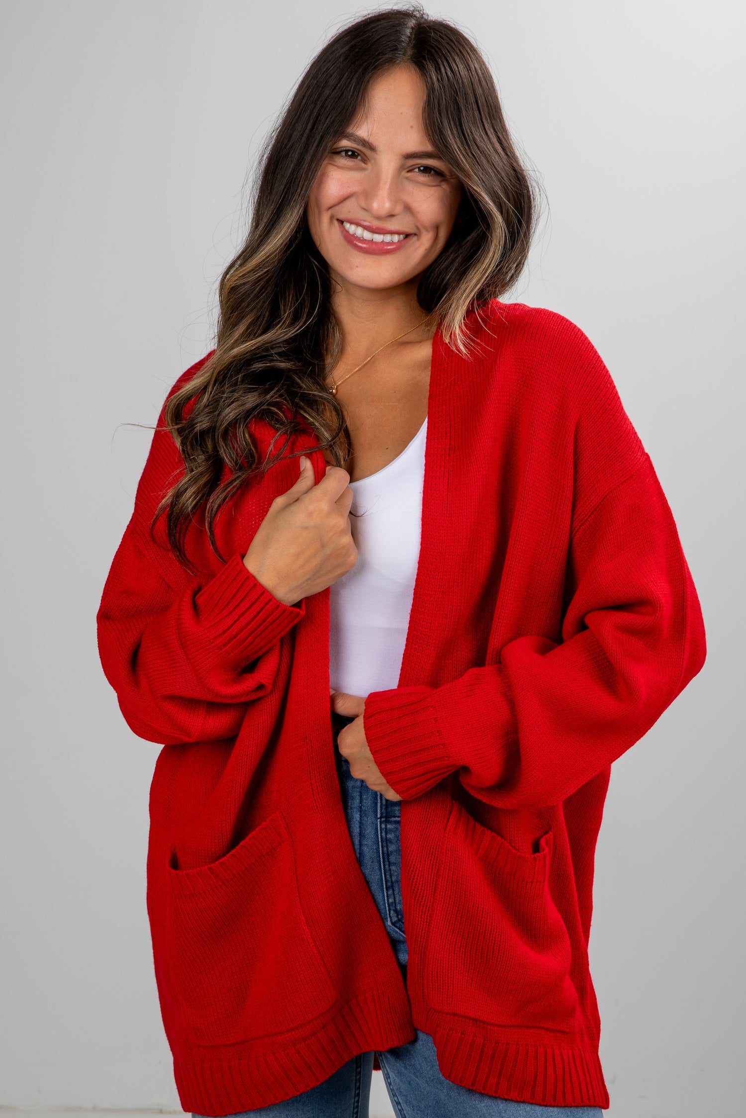 Woman wearing a red cardigan over a white top and blue jeans on a gray background