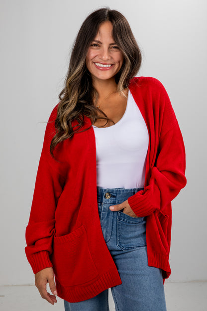 Woman wearing a red cardigan over a white top and blue jeans on a plain background