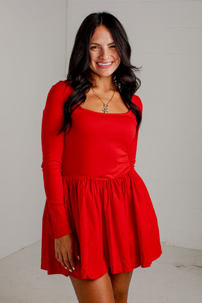 A woman modeling a bright red long-sleeve mini dress with a scoop neckline and a flared skirt, standing against a plain white wall.