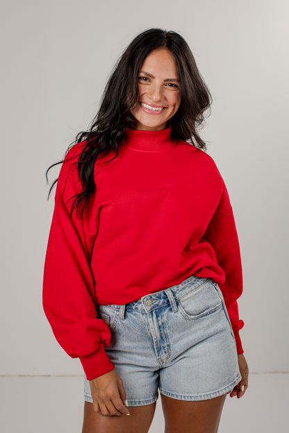 A woman with long dark hair wearing a bright red mock-neck sweatshirt and light-wash denim shorts, standing against a neutral light gray studio background.