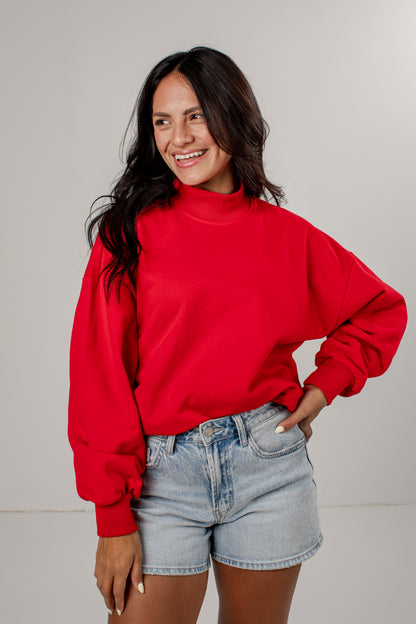 A woman wearing a bright red mock-neck sweatshirt and light-wash denim shorts, standing against a plain light gray studio background.