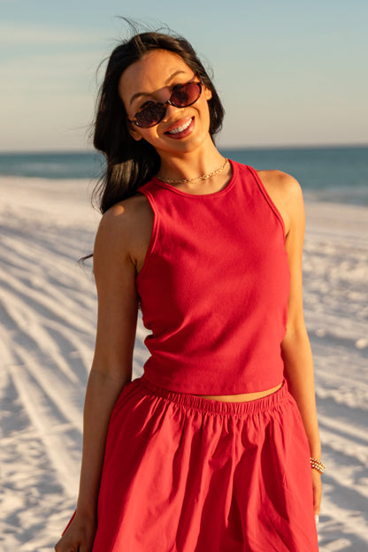 Woman in a red outfit standing on a sandy beach with sunglasses.