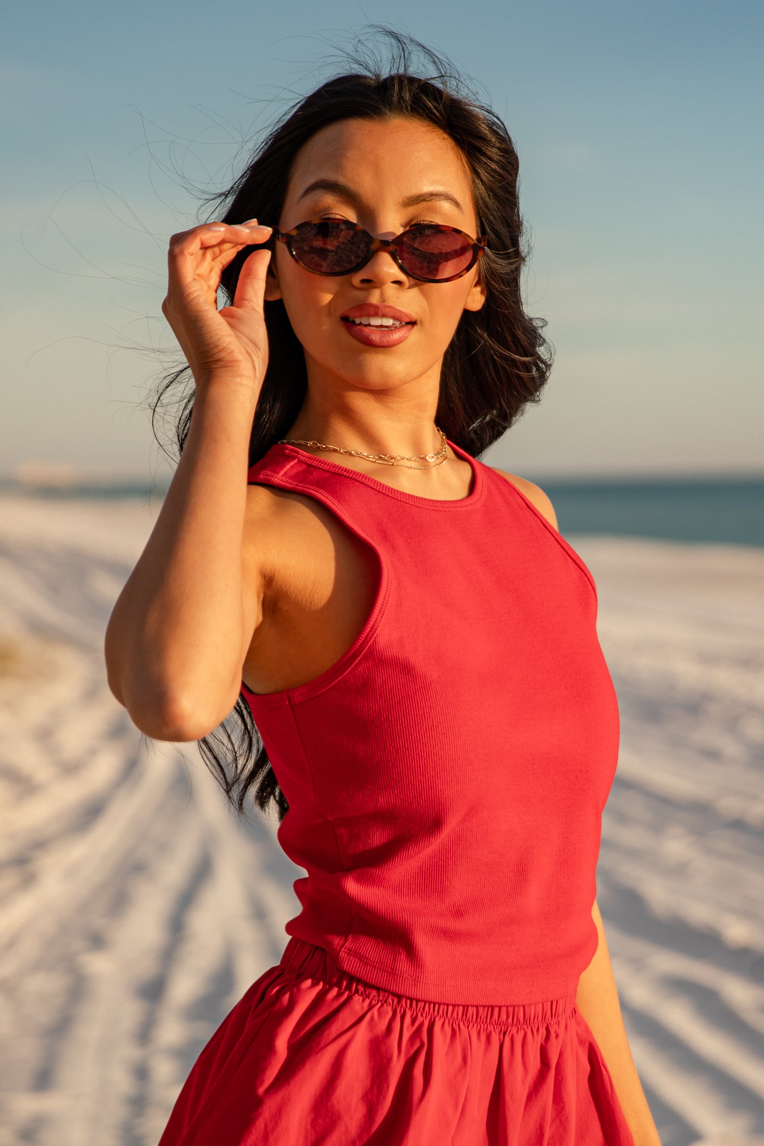 Woman in a red dress standing on a sandy beach with sunglasses, holding them up to her face.