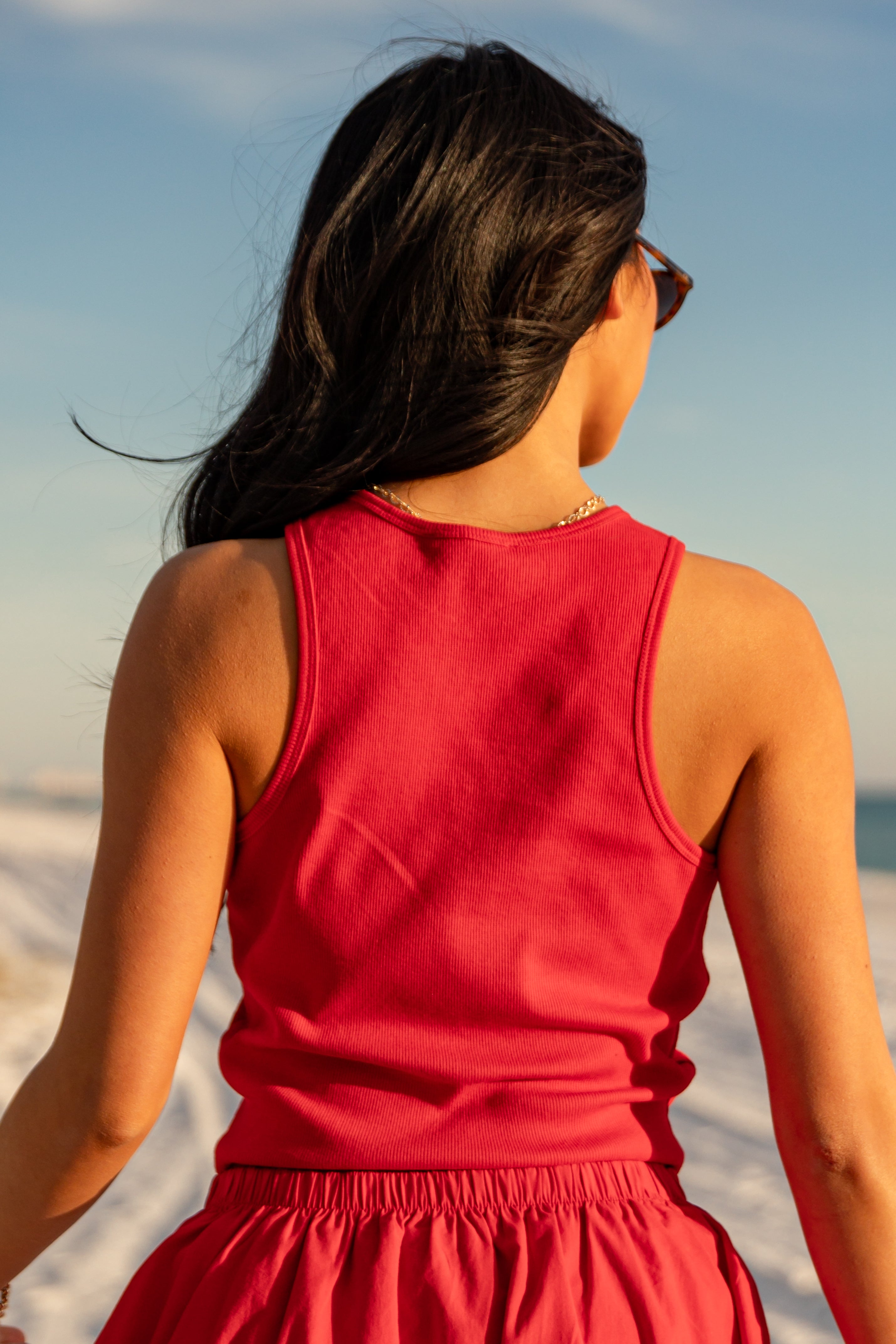 Woman in a red tank top and red skirt standing on a beach with a clear sky.