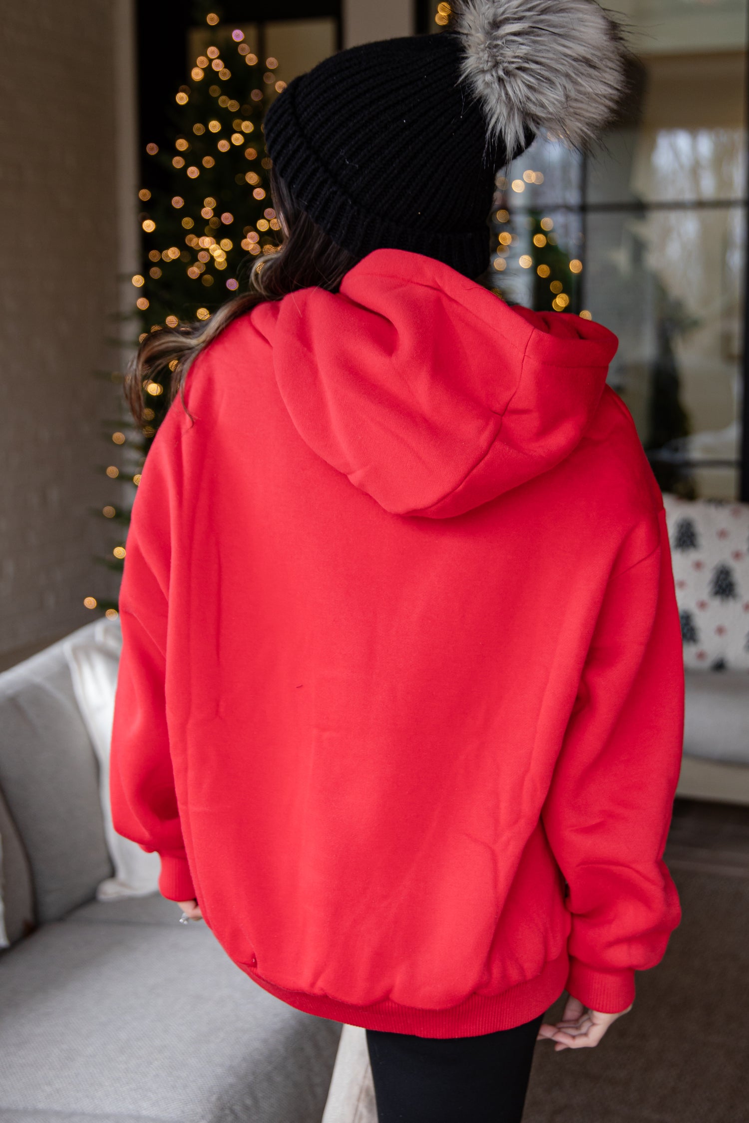 Person wearing a red hoodie with a black knit hat and gray fur pom-pom indoors.