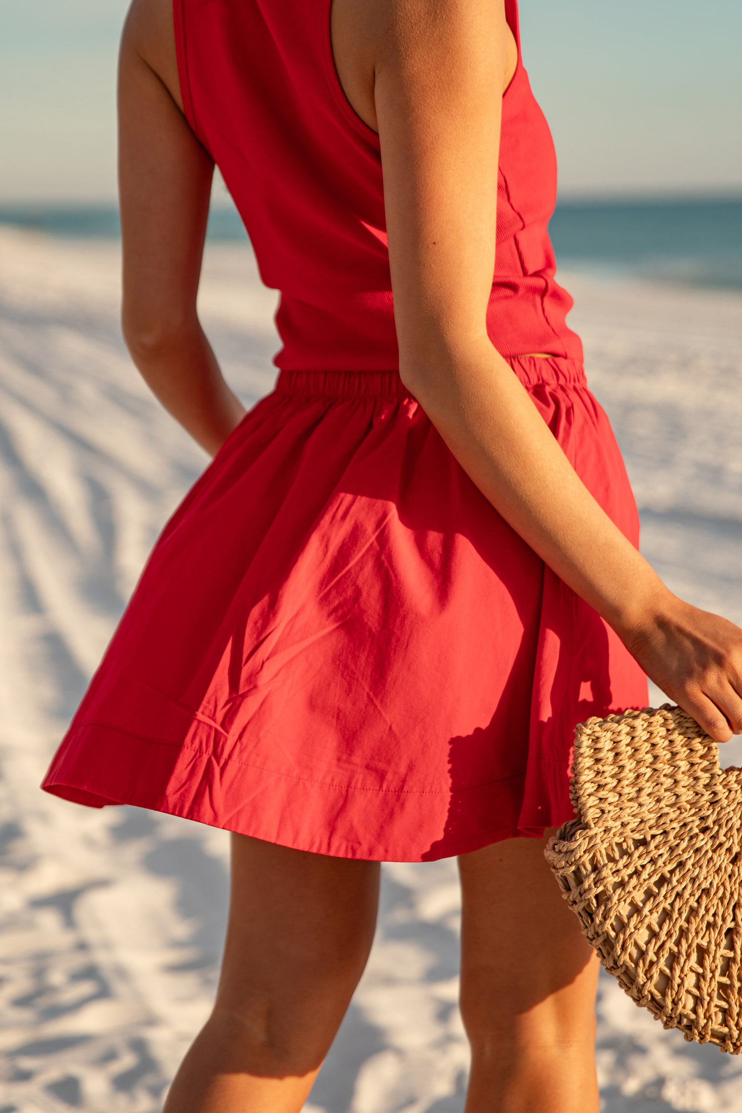 Person wearing a red dress on a beach holding a straw bag.