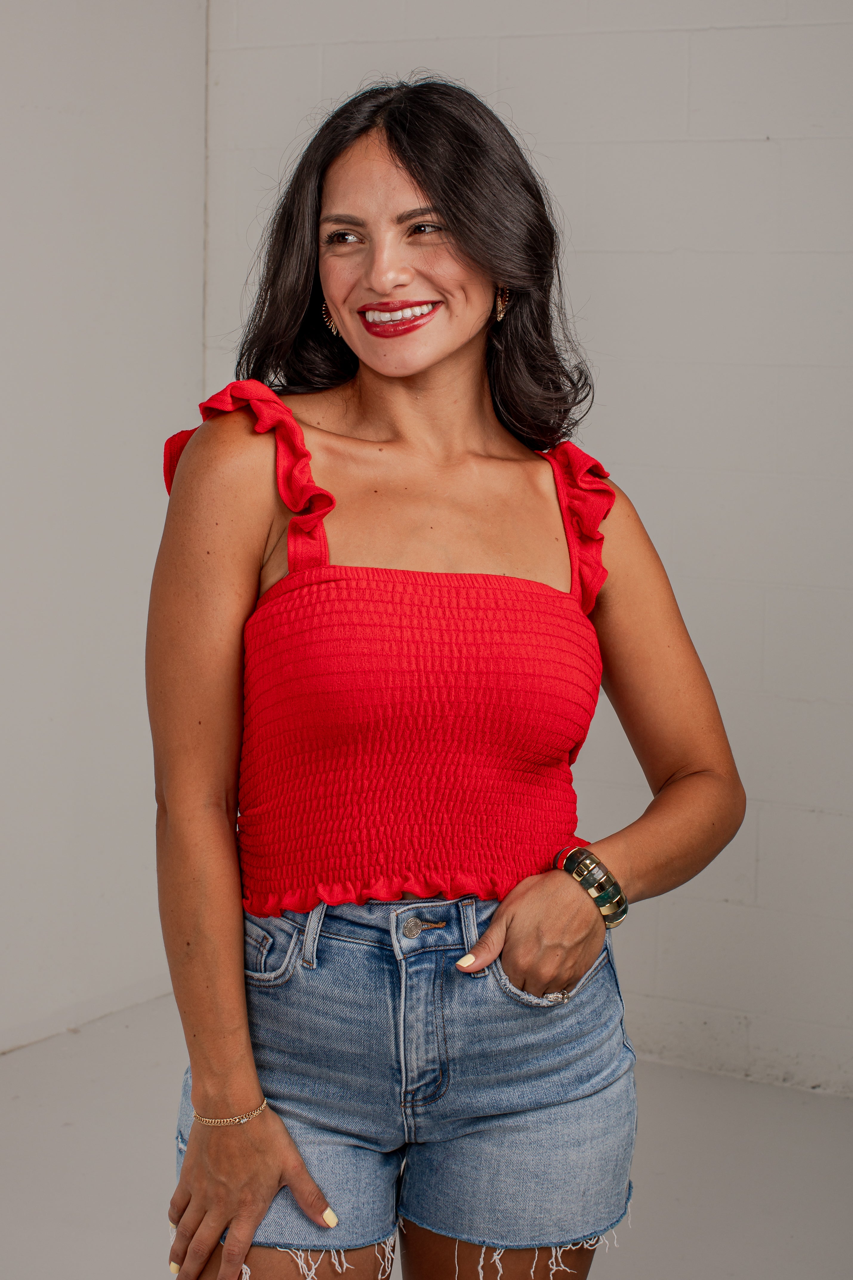 A woman wearing a bright red smocked camisole top with ruffled shoulder straps and light-wash denim shorts, standing against a light gray wall.