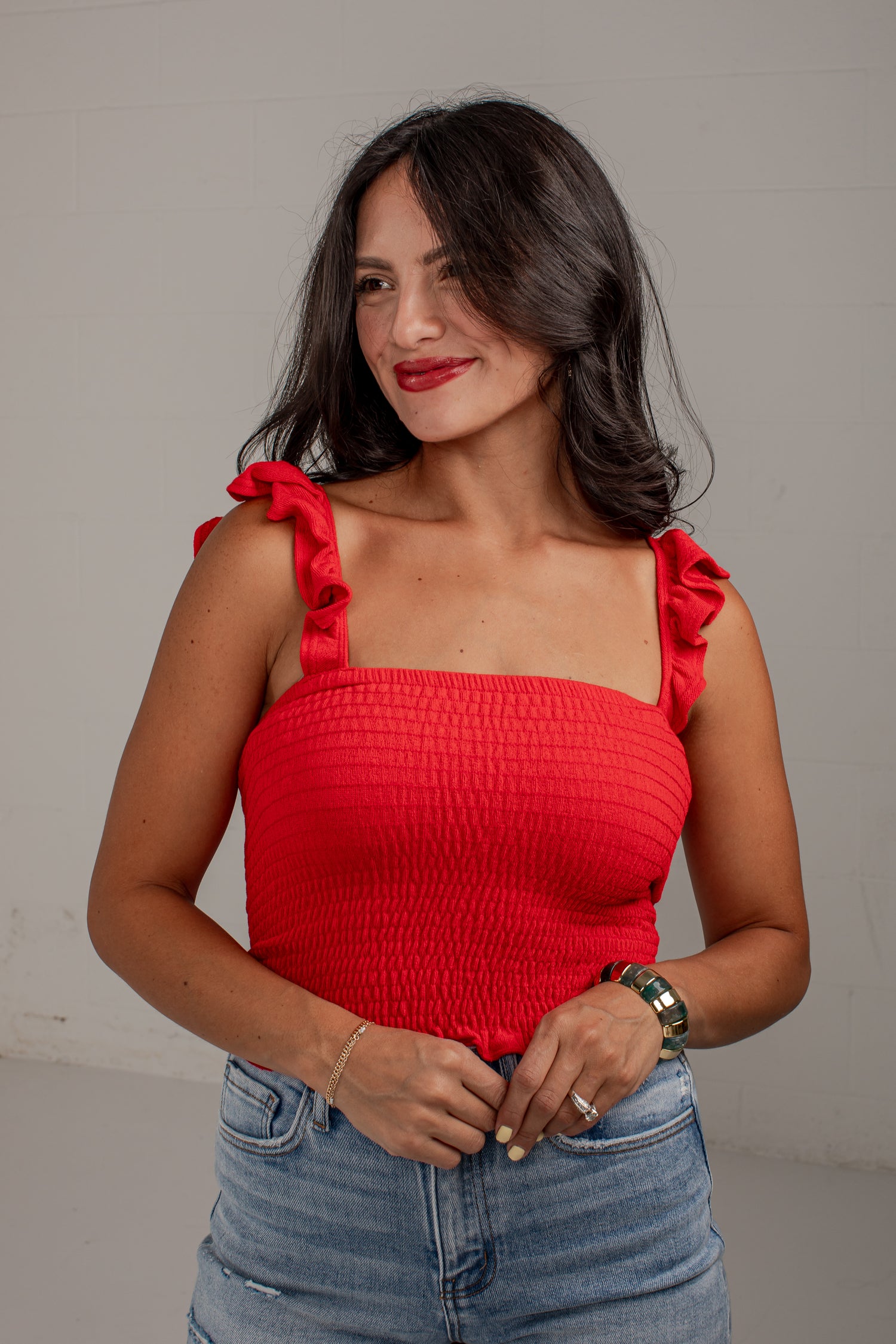 A woman wearing a bright red smocked crop top with ruffled shoulder straps and light-wash blue jeans, standing against a plain light gray wall.