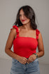 A woman wearing a bright red smocked crop top with ruffled shoulder straps and light-wash blue jeans, standing against a plain light gray wall.
