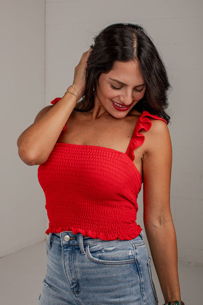 A woman wearing a bright red smocked crop top with ruffled shoulder straps and light wash blue jeans, posing against a neutral light gray wall.