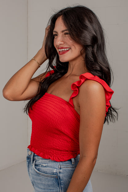 A woman wearing a bright red smocked one-shoulder ruffle top and blue jeans, posing in a studio setting against a light gray wall.
