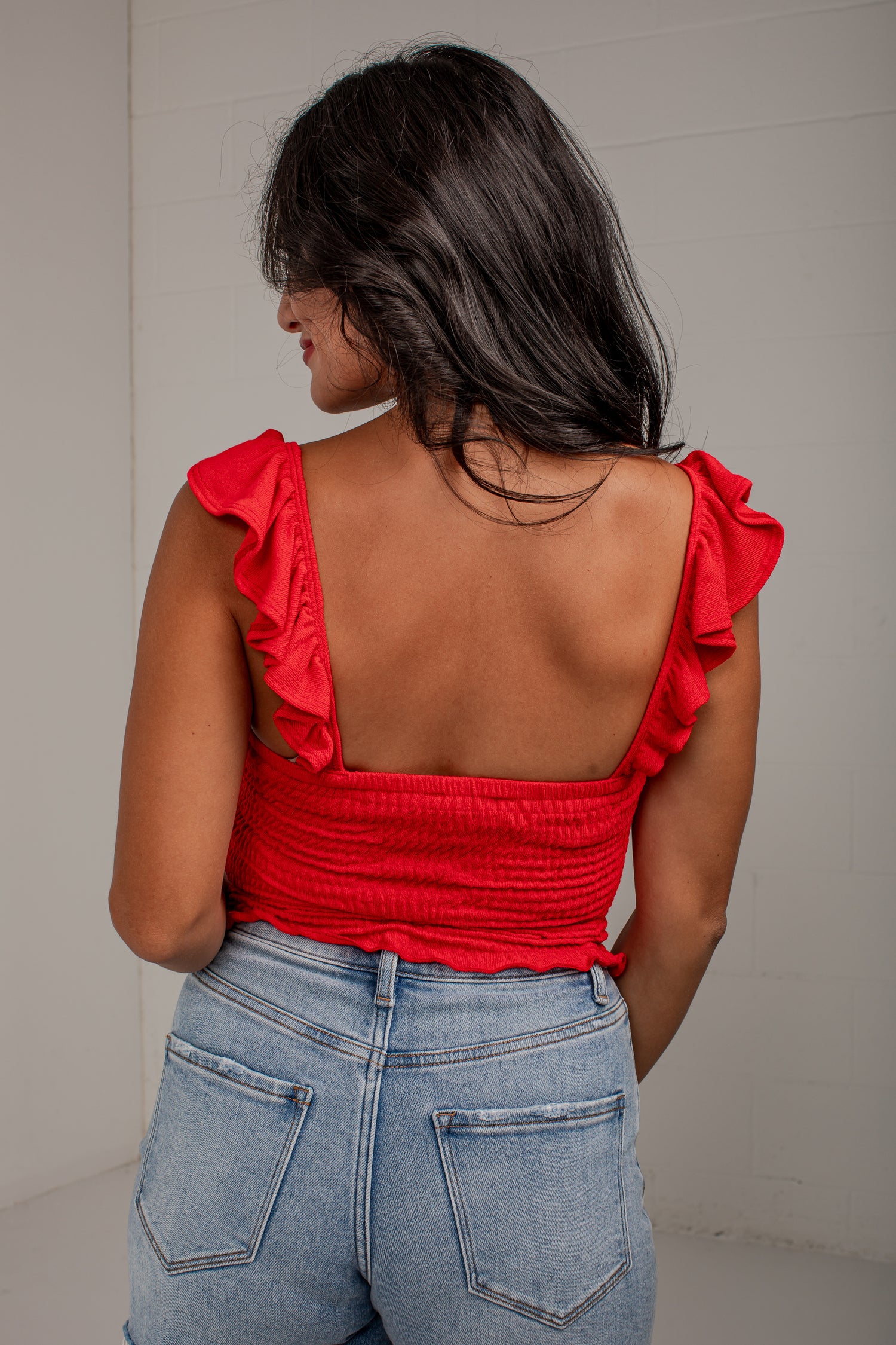 A woman shown from behind wearing a bright red ruffled strapless top with a smocked open back and light-wash blue jeans, standing against a plain light gray wall.