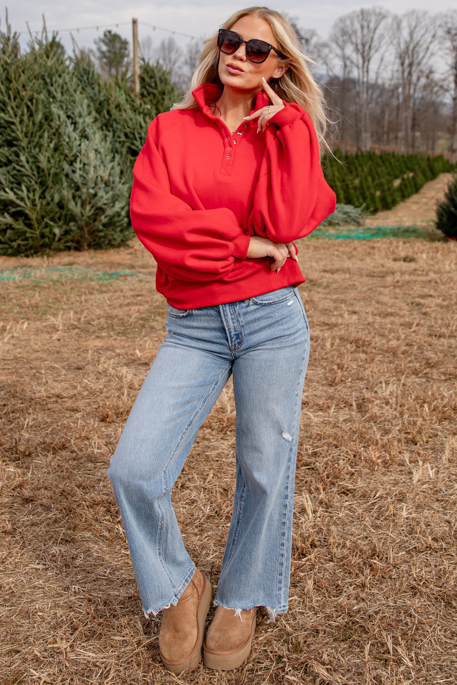 Woman in a red sweatshirt and blue jeans standing in a field with trees in the background