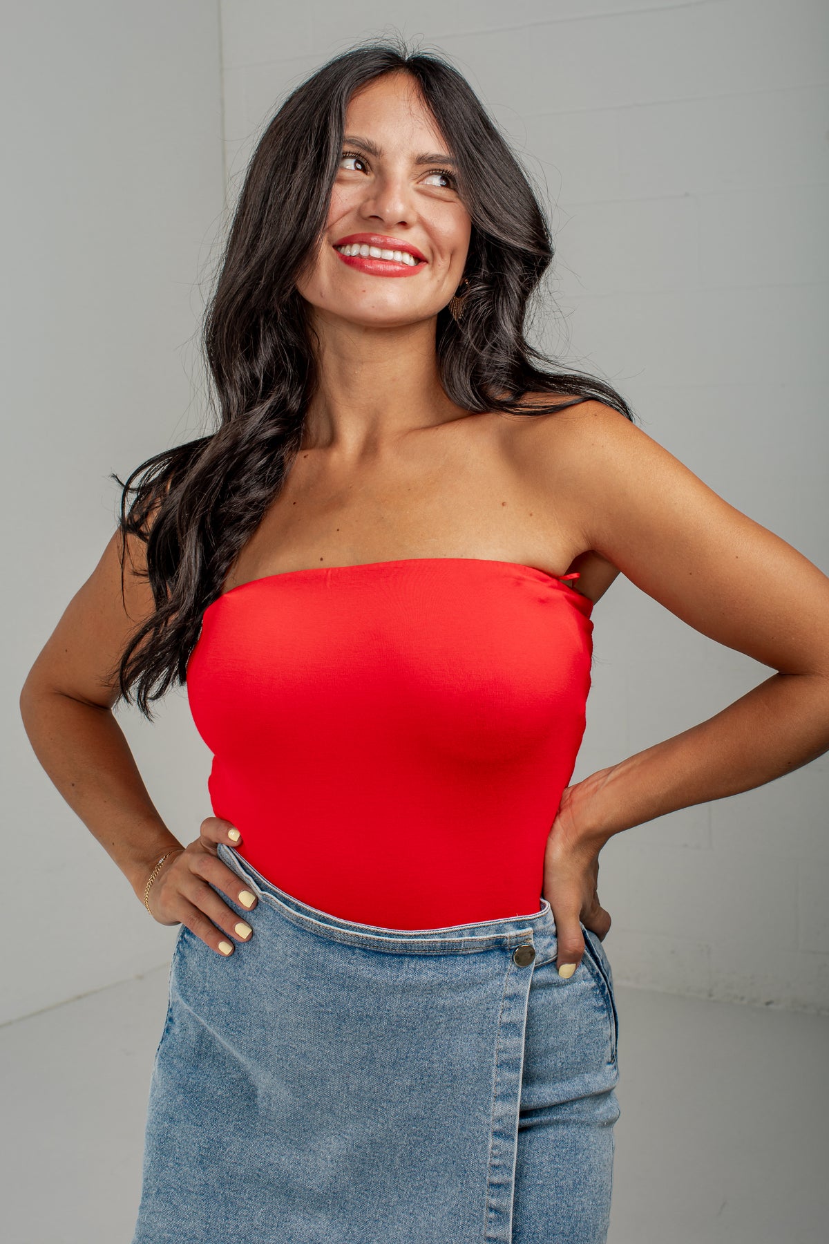 A woman wearing a bright red strapless tube top and a light blue denim skirt, posing against a light gray wall.