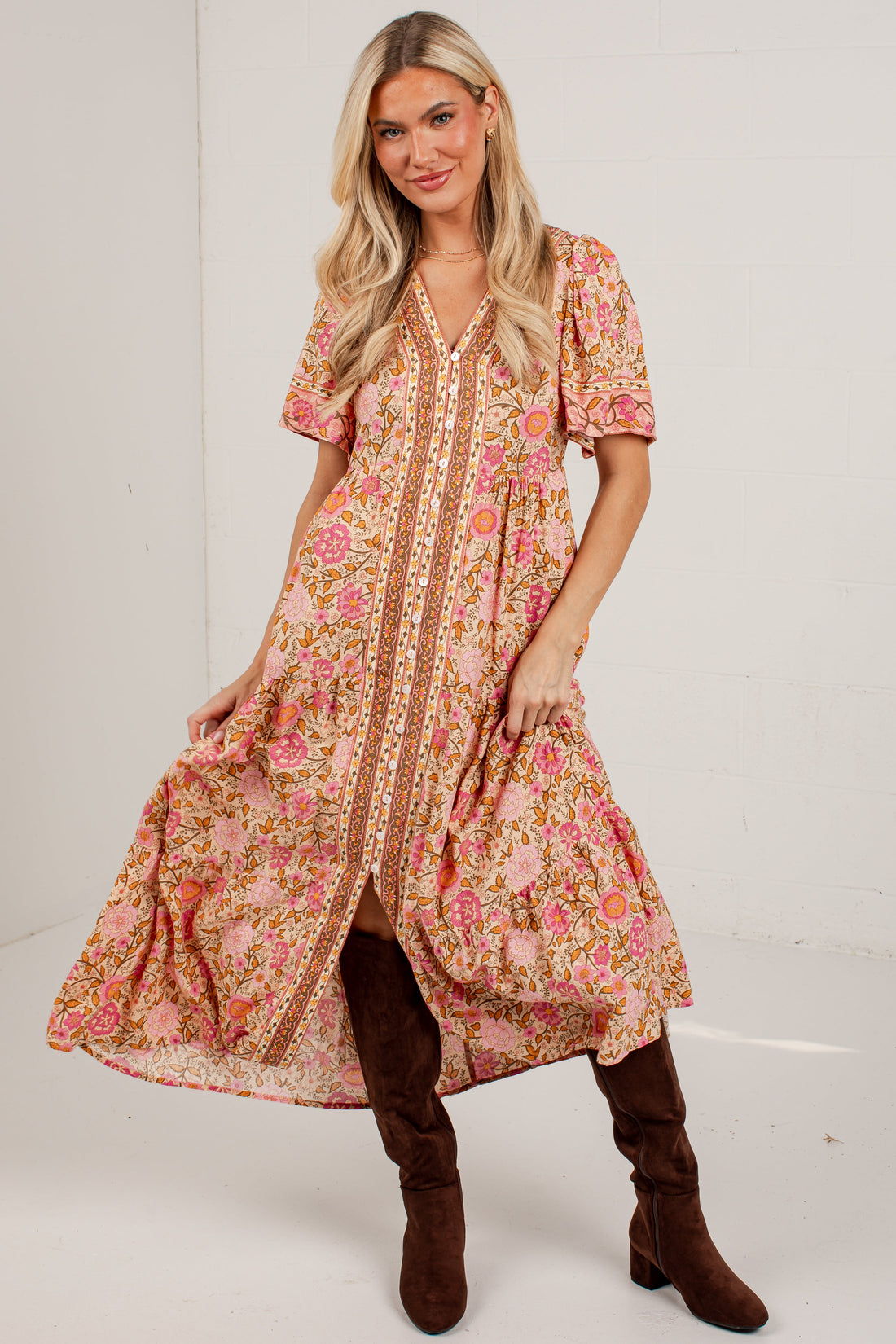 A woman modeling a bohemian-style midi dress with a pink and orange floral pattern, brown buttons, and puff sleeves, paired with brown suede boots against a white studio background.