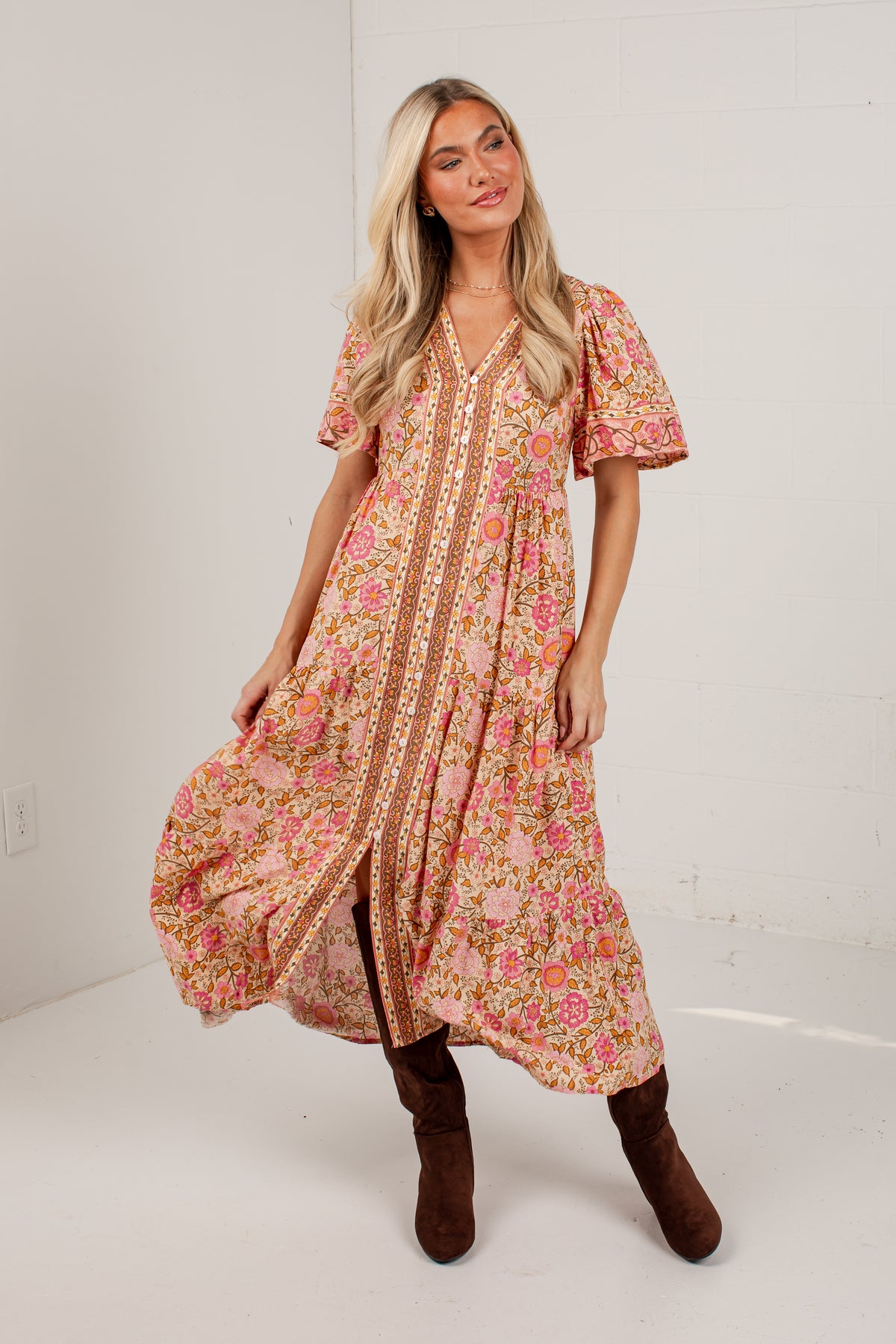 A woman modeling a short-sleeve bohemian-style maxi dress with a pink and orange floral pattern and a gold patterned center panel, paired with brown boots, in a white studio setting.