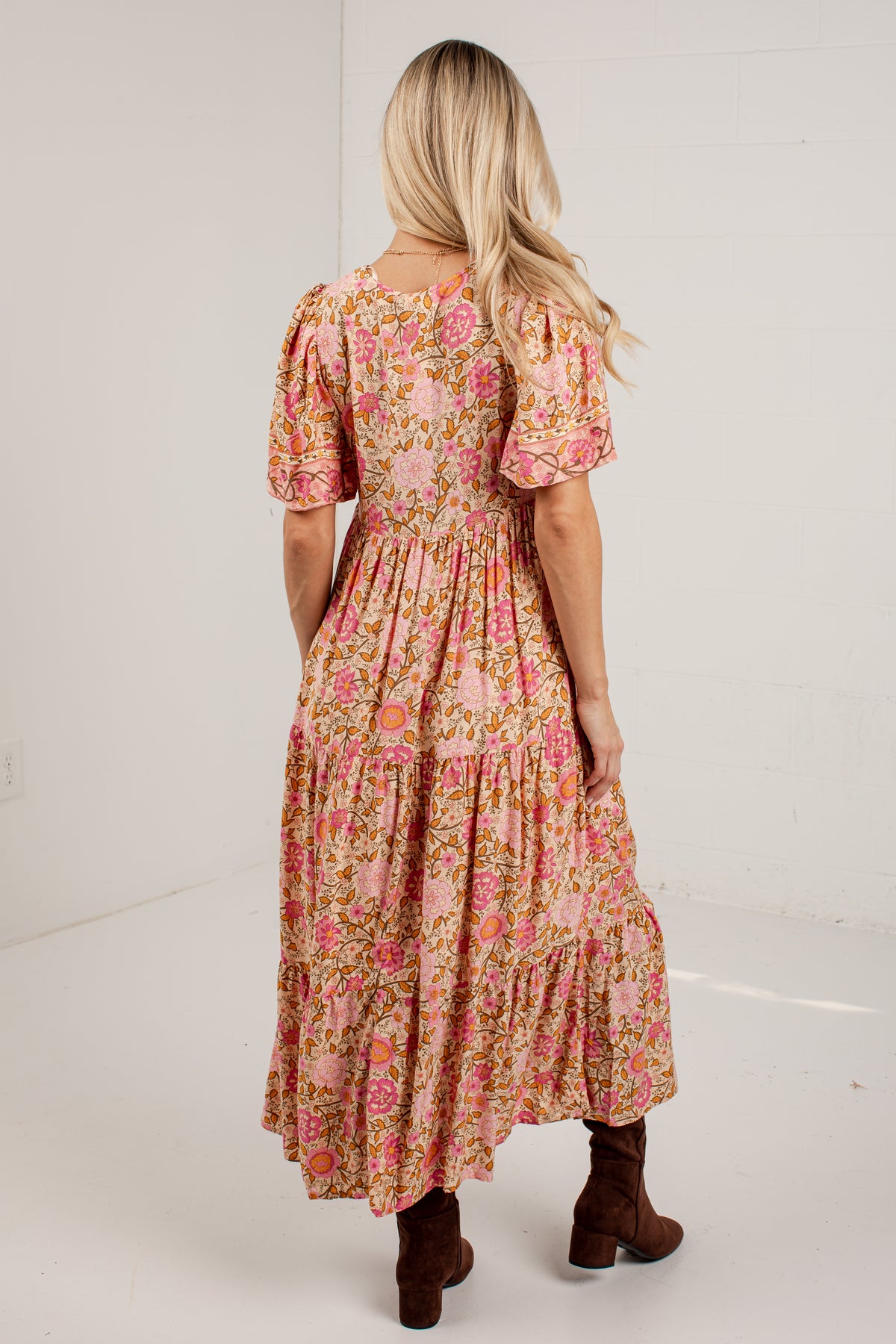 A woman viewed from behind wearing a pink floral tiered midi dress with puff sleeves and brown ankle boots, standing against a white studio wall.