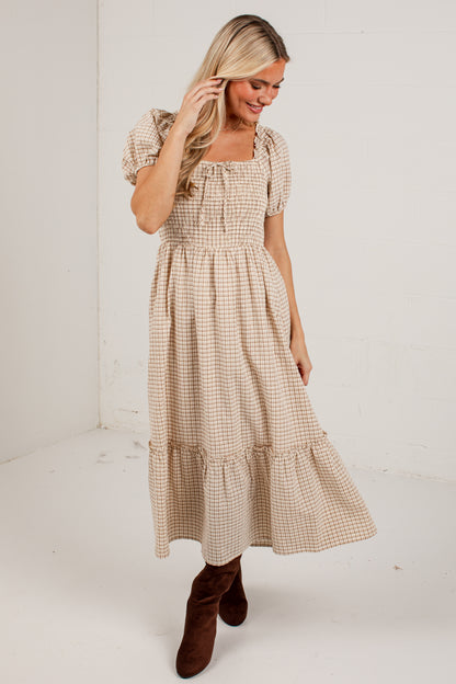 A woman wearing a beige and brown gingham midi dress with puff sleeves and a smocked bodice, paired with brown suede boots, standing in a white studio corner.
