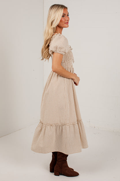 A woman modeling a beige and white gingham tiered midi dress with puff sleeves, shown from a side-back angle against a white studio background.