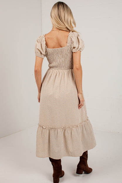 A woman viewed from the back wearing a beige and white gingham midi dress with puff sleeves and a smocked bodice, paired with brown ankle boots, against a white studio background.