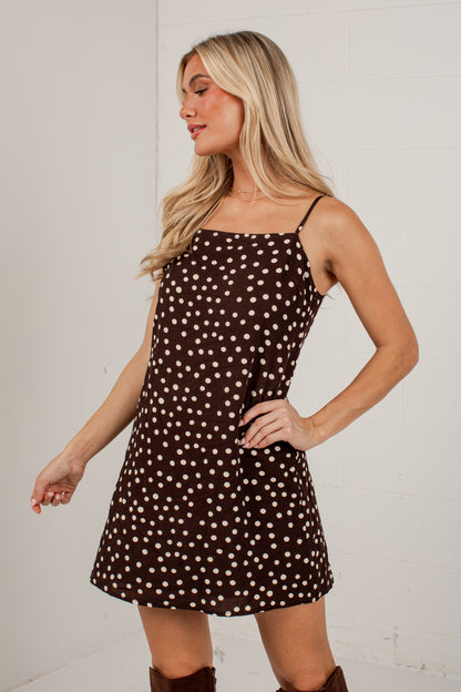 A blonde woman modeling a brown mini dress with a white polka dot pattern, featuring thin spaghetti straps and a square neckline, posed against a white brick wall.