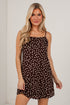 A woman modeling a brown spaghetti-strap mini dress with a white polka dot pattern against a white brick wall background.