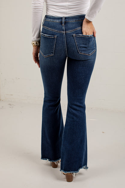 A person shown from the back wearing dark blue flare jeans with a frayed hem, paired with a white long-sleeve top and brown heeled shoes, standing against a plain off-white wall.