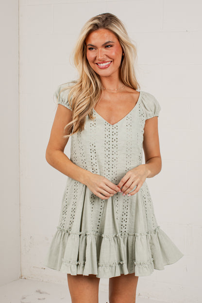 A woman modeling a sage green eyelet mini dress with puff sleeves and a tiered ruffle hem, standing against a white brick wall.